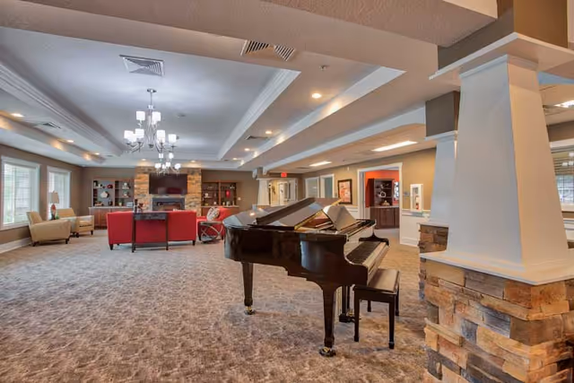 Spacious senior living common area with a grand piano in the foreground, comfortable seating including red sofas and beige armchairs, a stone fireplace with a mounted TV, and large windows letting in natural light.