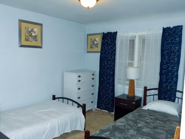 A small bedroom with two single beds, one with a white bedspread and the other with a gray bedspread. Between the beds is a dark wooden nightstand with a lamp on it. There is a white chest of drawers against the wall and two framed pictures hanging above it. The window has white blinds and dark blue curtains with a swirl pattern. The walls are painted light blue and the floor is carpeted.