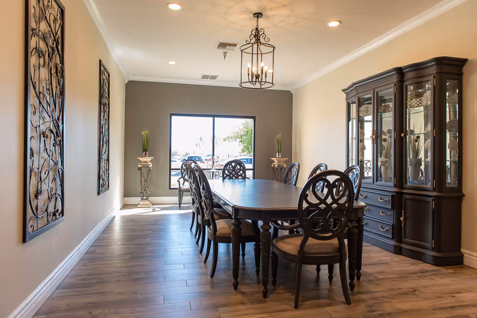 A well-lit dining room with a long dark wooden table surrounded by eight matching chairs. The room features a large window at the far end letting in natural light, two decorative plant stands with tall plants on either side of the window, two ornate wall hangings on the left wall, a chandelier hanging from the ceiling, and a large dark wooden china cabinet on the right side filled with decorative items.
