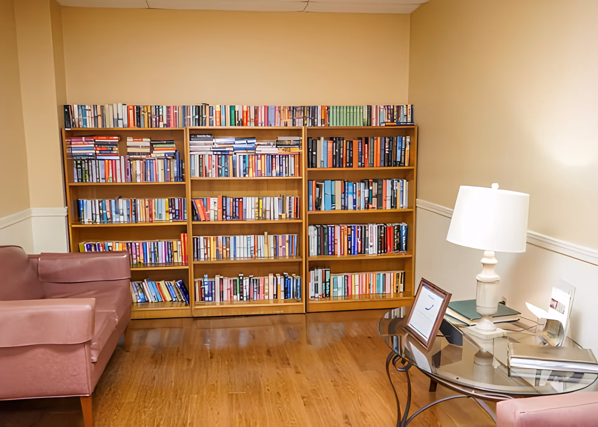 A cozy room with wooden bookshelves filled with books against a beige wall. There is a pink armchair on the left and a glass-top table on the right with a white table lamp, some books, and a framed picture. The floor is wooden.