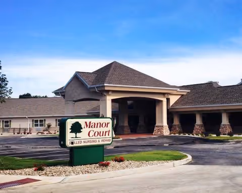 Exterior view of a single-story nursing and rehab facility named Manor Court, featuring a covered entrance with stone pillars, a parking area, and a sign in front displaying the facility's name and services.