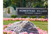 Entrance sign reading "Homestead Village Retirement Community" set among pink flowers, grass, a large rock, and trees.
