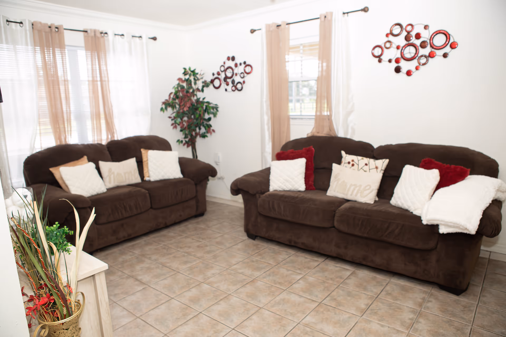 Bright living room with two brown sofas, decorative throw pillows, wall art, and tiled floor.