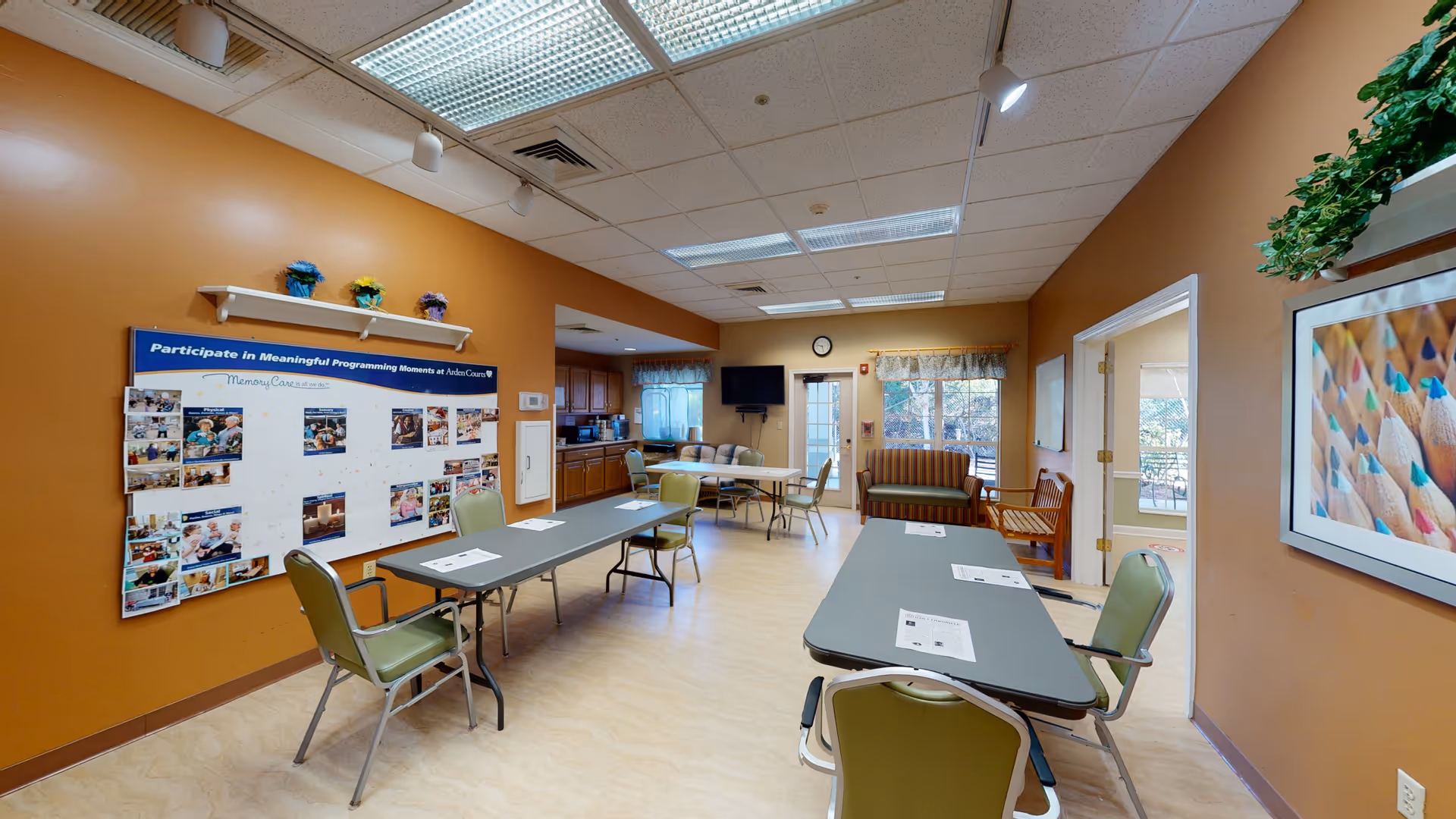 A community room with brown walls and a light-colored floor featuring two long tables with green chairs around them. On the left wall, there is a bulletin board titled 'Participate in Meaningful Programming Moments at Arden Courts' with photos and notes. The back of the room has a small kitchenette area with cabinets and a window. There is a wall-mounted TV and a clock above a door leading outside. On the right wall, there is a framed picture of colored pencils and a small plant on a shelf. The room is well-lit with ceiling lights.