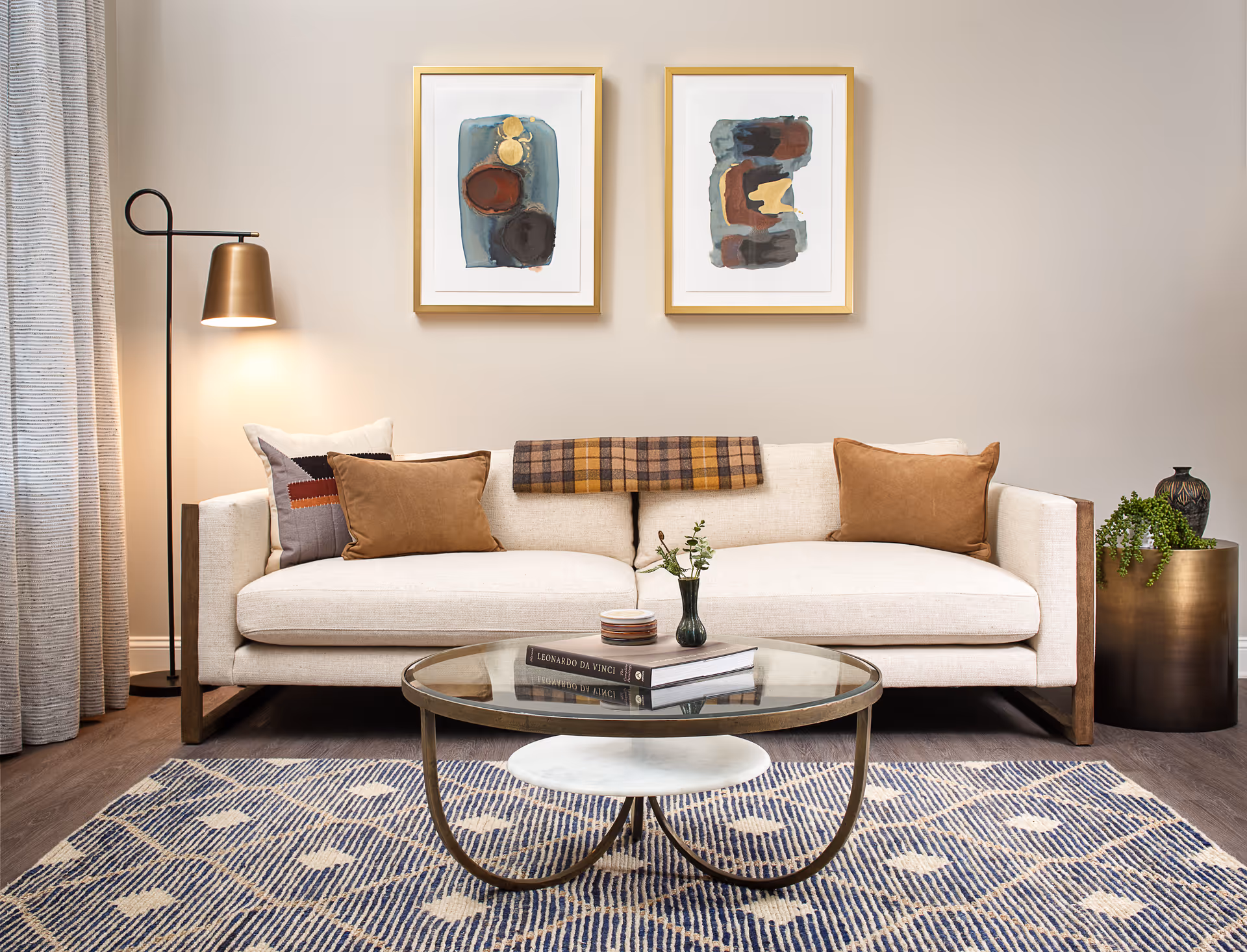 A modern living room with a beige sofa adorned with brown and patterned cushions, a plaid throw draped over the back, a round glass coffee table with books and a small vase, a floor lamp with a brass shade, two abstract paintings in gold frames on the wall, a patterned rug, and a brass planter with a green plant.
