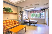 A bright and spacious patient room in a care facility featuring a hospital bed near a large window with a floral valance, a plaid orange and yellow couch, a wooden coffee table, and a wall-mounted television. There is also a shelf with green plants and medical equipment mounted on the wall.