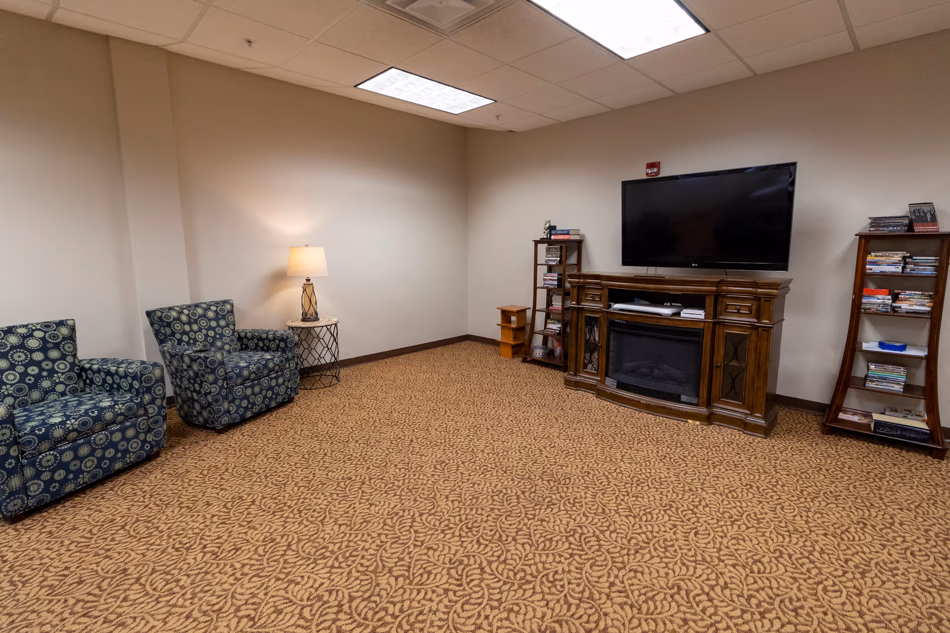 A cozy living room area with two patterned armchairs, a small round side table with a lamp, a large flat-screen TV mounted on a wooden entertainment center with a built-in electric fireplace, and two wooden shelves filled with books and DVDs. The room has beige walls and a patterned carpet.