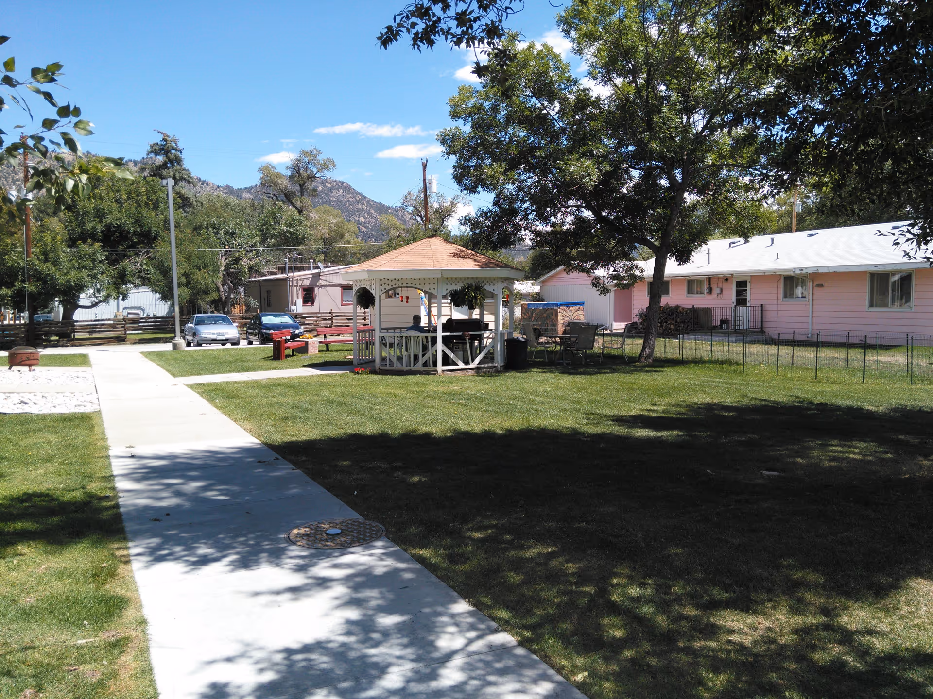 Outdoor area of a senior living facility with a concrete walkway leading to a white gazebo. The gazebo has hanging plants and people sitting inside. There are trees providing shade, a pink building in the background, parked cars, and mountains visible in the distance under a clear blue sky.
