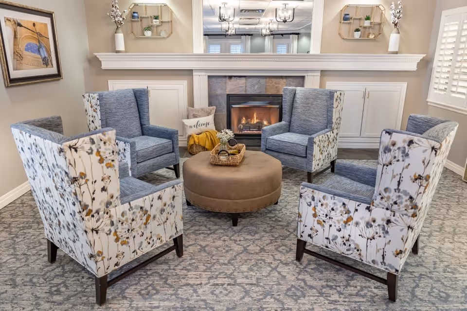 A cozy sitting area with four armchairs arranged around a round ottoman in front of a lit fireplace. The chairs have a floral pattern on the outside and a solid blue fabric on the inside. The room features a patterned carpet, a large mirror above the fireplace, decorative shelves with plants, and a framed picture on the wall.