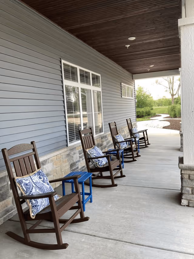 A covered outdoor porch area with four wooden rocking chairs, each with a blue and white patterned cushion. There are small blue side tables between the chairs. The porch has a stone and blue siding exterior wall, and a wooden ceiling. A curved pathway and greenery are visible in the background.
