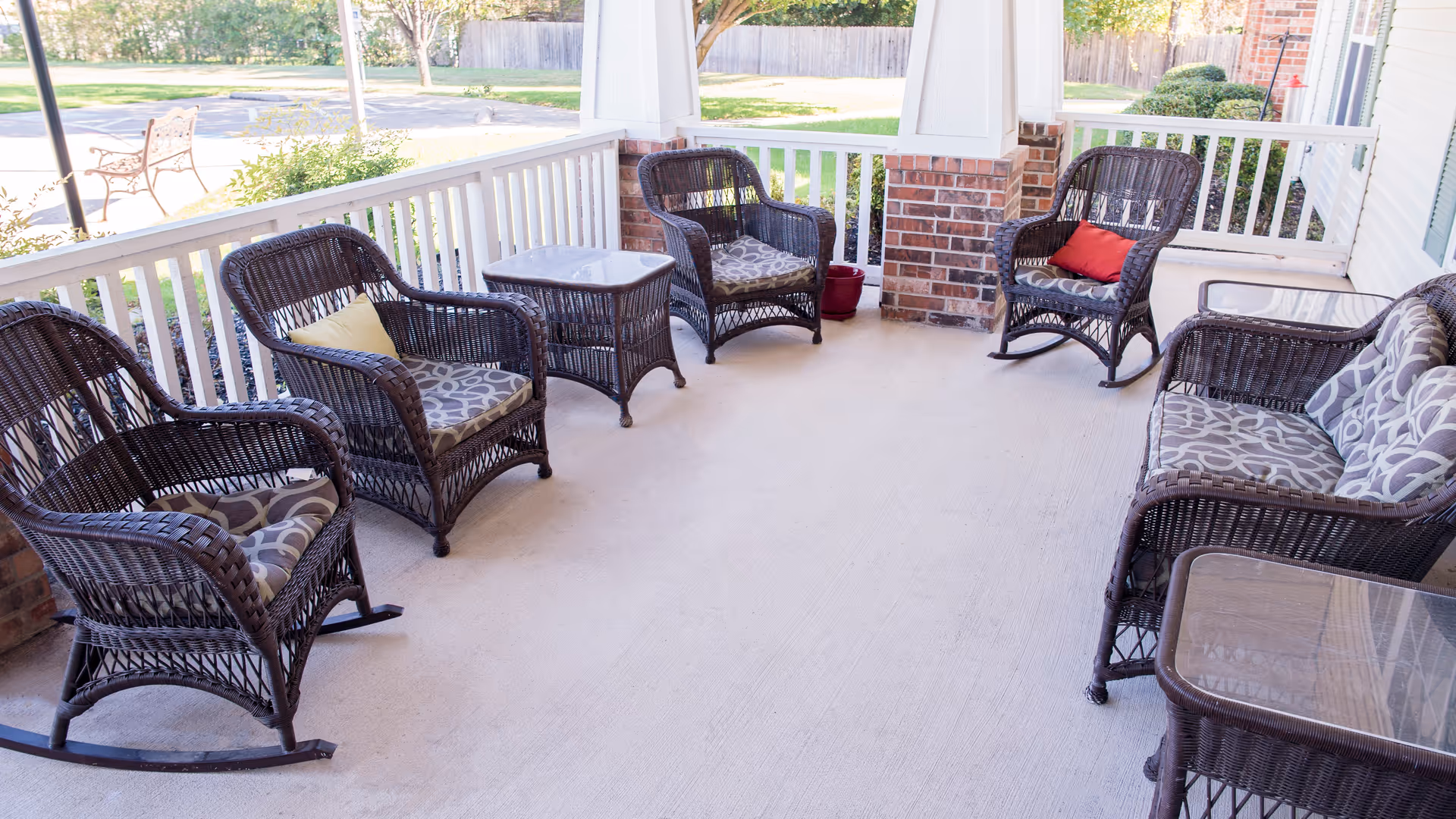 A covered outdoor porch area with several dark wicker chairs and a loveseat, all with patterned cushions. There are two small glass-top wicker tables, a white railing, brick pillars, and a view of a green lawn and trees beyond the porch.