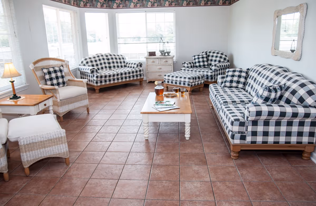A bright living room with large windows and tiled floor featuring a checkered pattern sofa set including a couch, loveseat, armchair, and ottoman. There is a wooden coffee table in the center with a glass pitcher, two glasses, and some magazines. A wicker chair with a cushion and a small wooden side table with a lamp are on the left side. A mirror hangs on the wall above the sofa on the right.