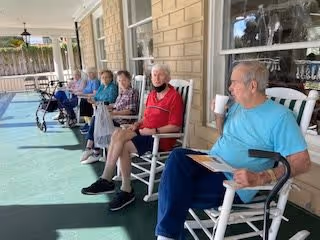 A group of elderly people sitting on rocking chairs on a covered porch outside a building. They appear relaxed, some holding drinks or reading materials, with a walker visible next to one person. The porch has a green floor and beige brick walls with windows.