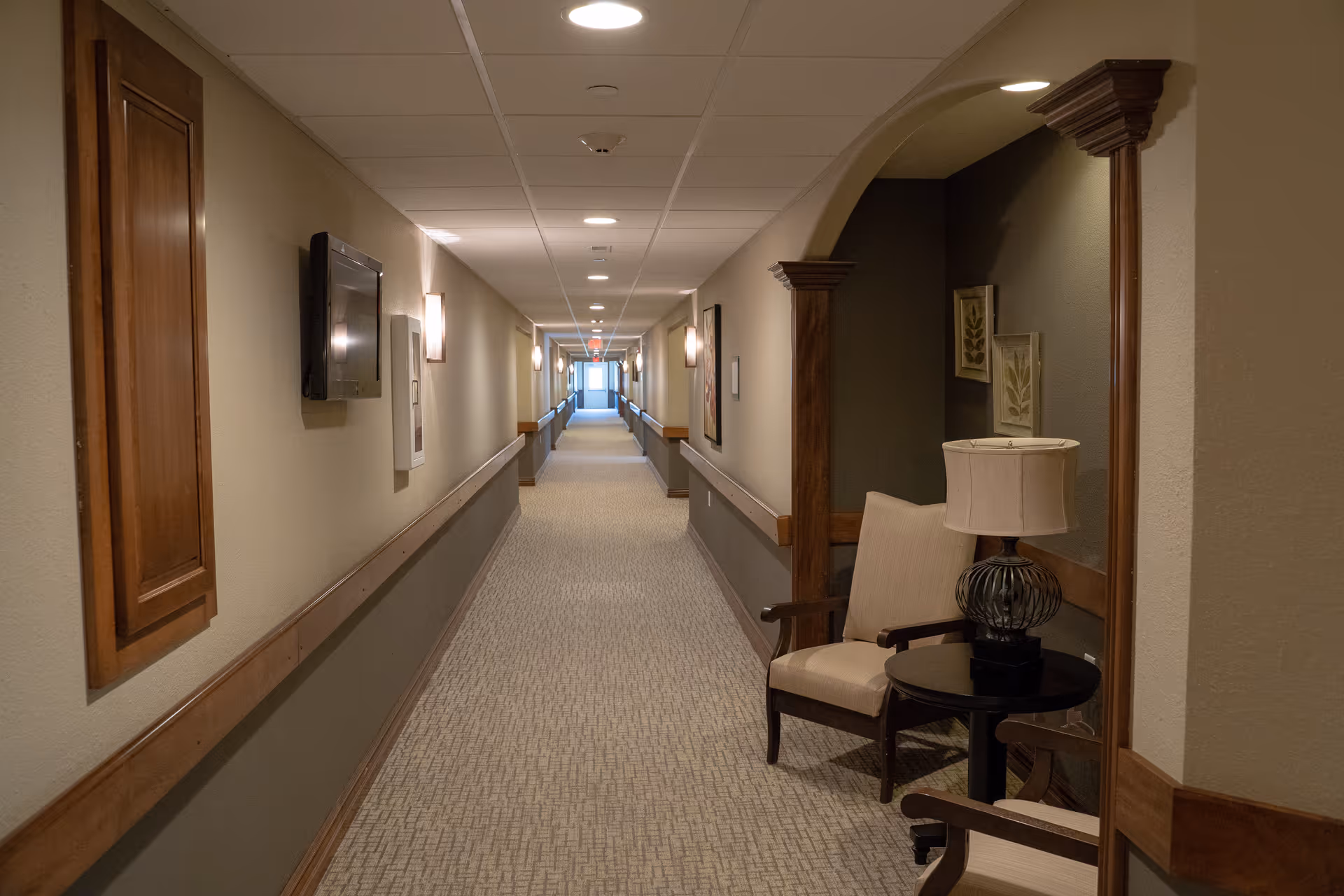 A long, well-lit hallway in a senior living facility with beige walls and carpeted floor. The hallway has wooden handrails on both sides and several wall-mounted lights. On the right side near the foreground, there is a small seating area with two beige armchairs and a round table with a lamp. Decorative framed artwork is hung on the walls.