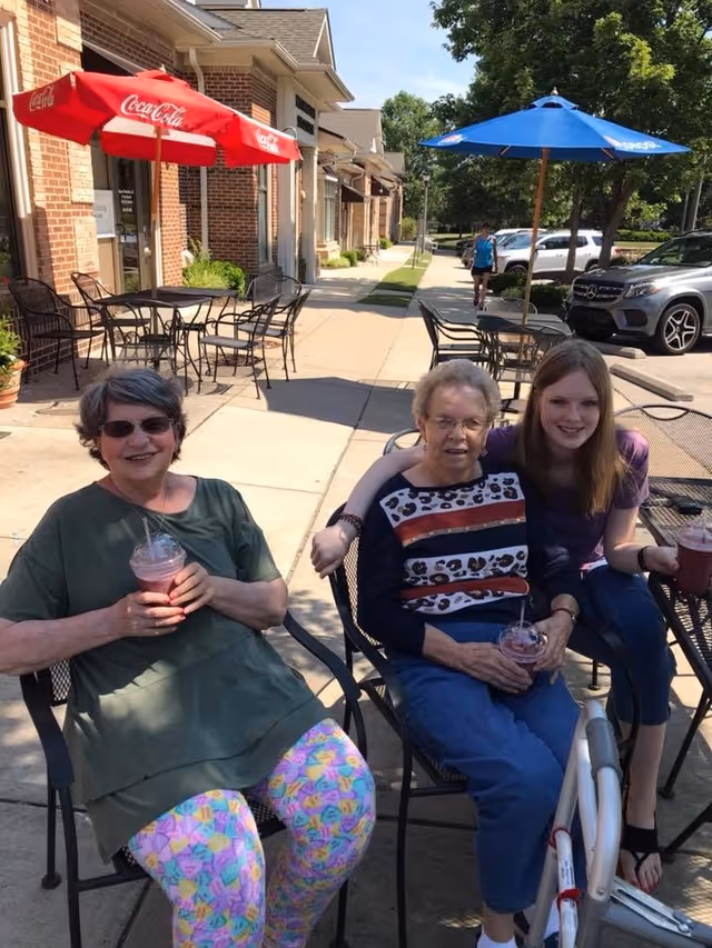 Three women sitting outdoors at a patio table with metal chairs, enjoying drinks. Two older women and one younger woman are smiling at the camera. There are red and blue Coca-Cola umbrellas providing shade, and a sidewalk with parked cars and trees in the background.
