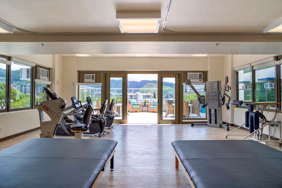 A bright fitness room with exercise equipment including stationary bikes and weight machines, two padded therapy tables in the foreground, large windows on three sides, and glass doors leading to an outdoor patio with seating and a view of green hills in the background.