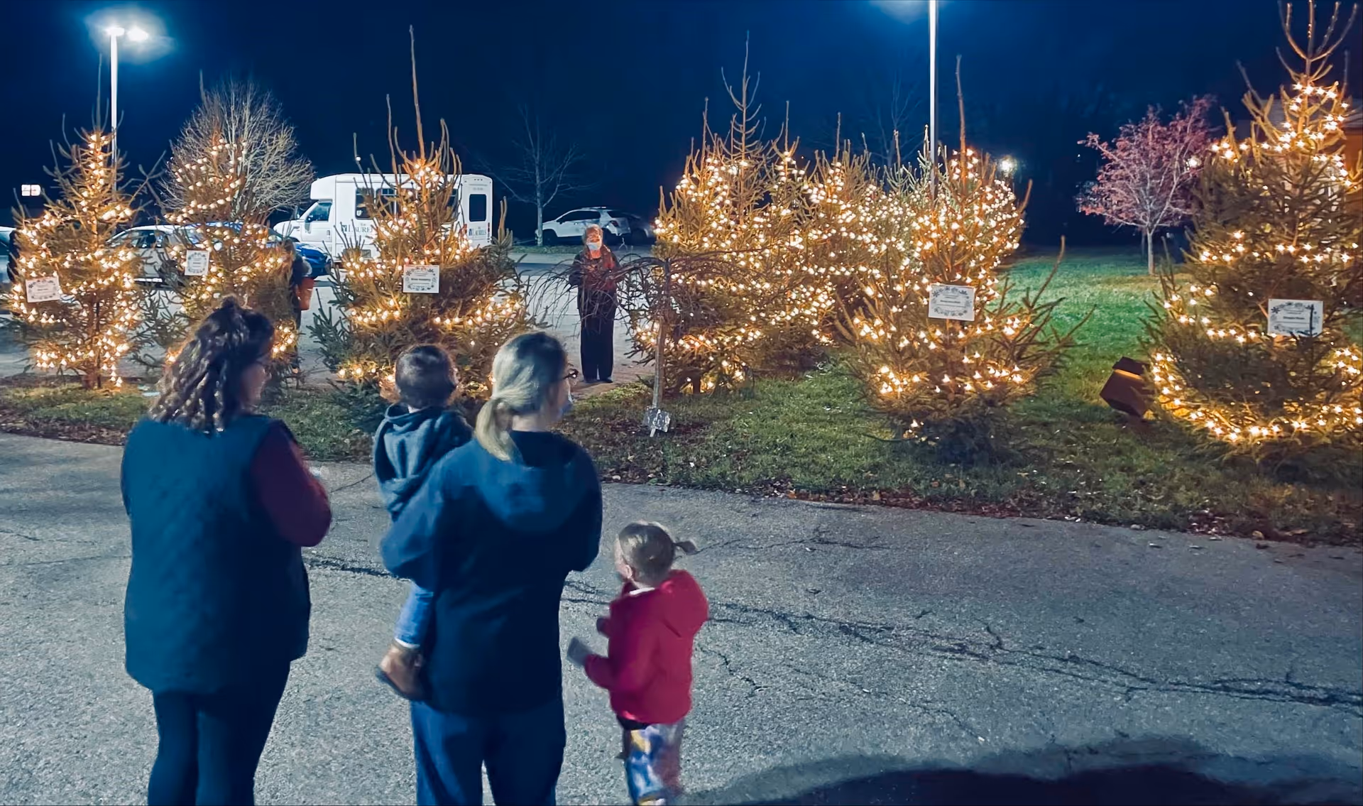 A small group of adults and children stand in a parking lot at night looking at rows of Christmas trees wrapped in string lights.