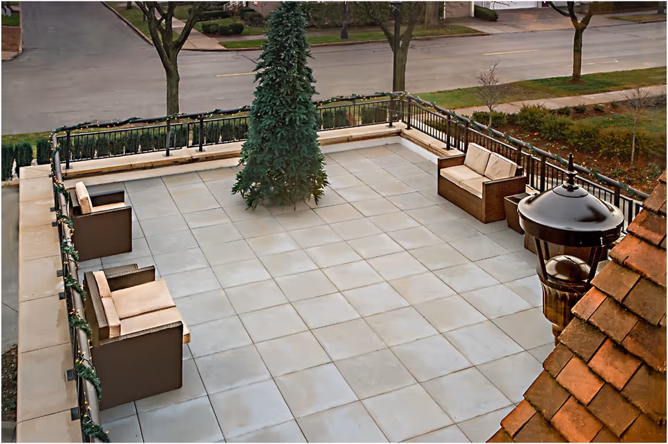 Outdoor patio area with tiled flooring, featuring a decorated Christmas tree in the center, surrounded by cushioned wicker chairs and sofas. The patio is enclosed by a black metal railing adorned with garland and lights. A street and some trees are visible in the background.