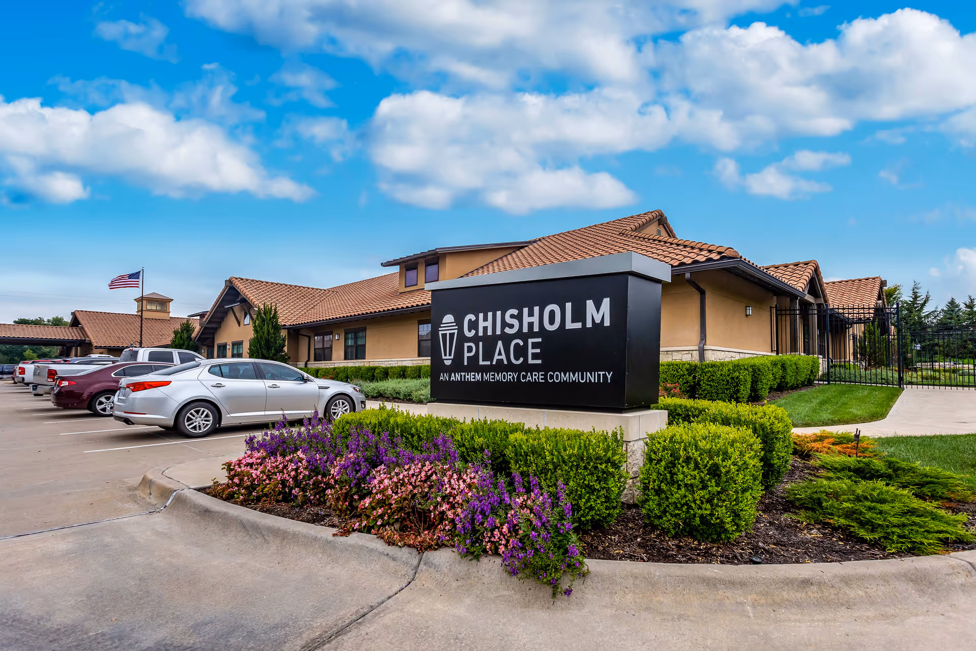 Exterior view of Chisholm Place, an Anthem Memory Care Community, showing a single-story building with a tiled roof, a parking lot with several cars, an American flag, and landscaped bushes and flowers in the foreground under a partly cloudy sky.