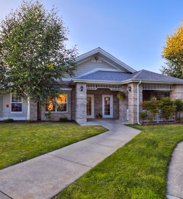 Front exterior view of a single-story building with a covered entrance, stone and siding facade, surrounded by green grass and trees under a clear blue sky.