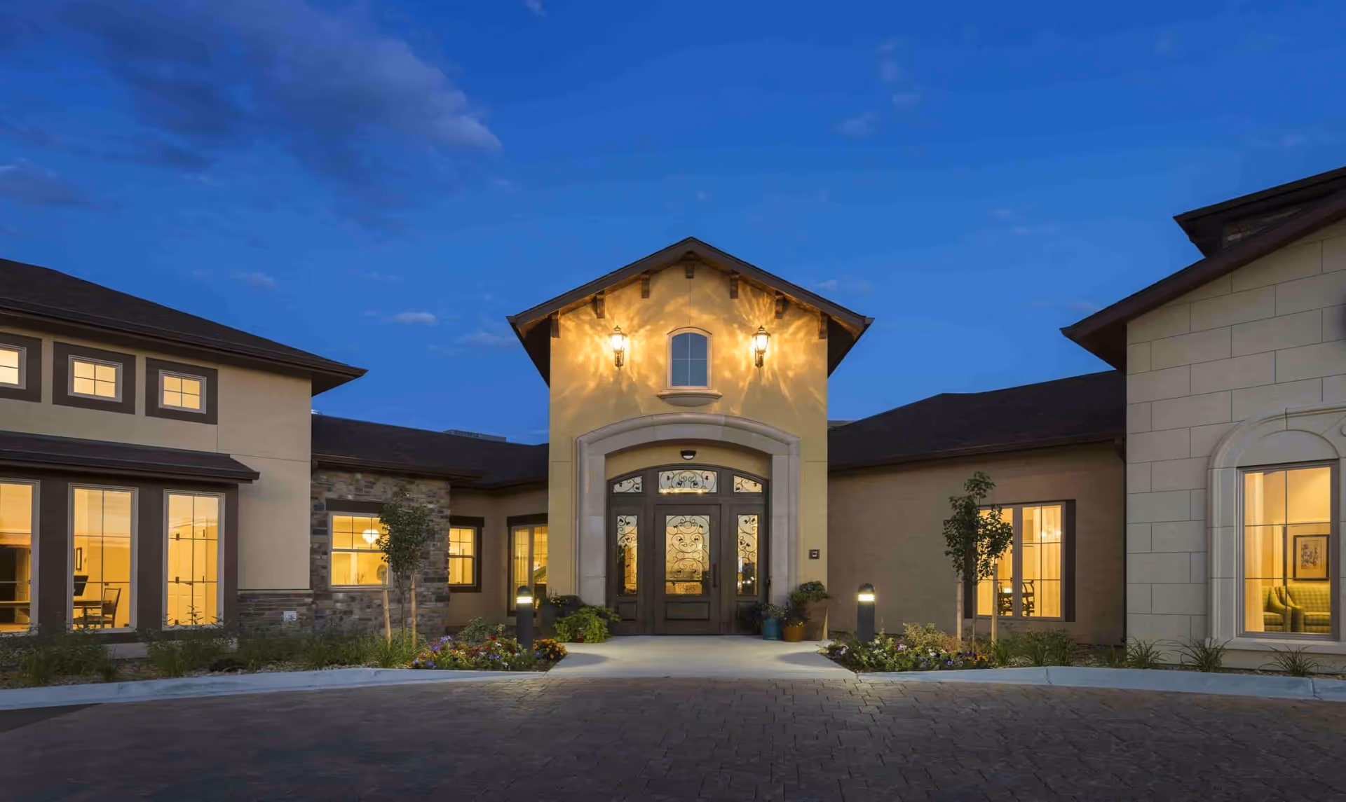 Evening view of the entrance to a senior living facility with warm exterior lighting, large decorative double doors, and windows showing interior rooms with lights on. The building has a mix of stone and stucco walls with a paved driveway and landscaped flower beds in front.