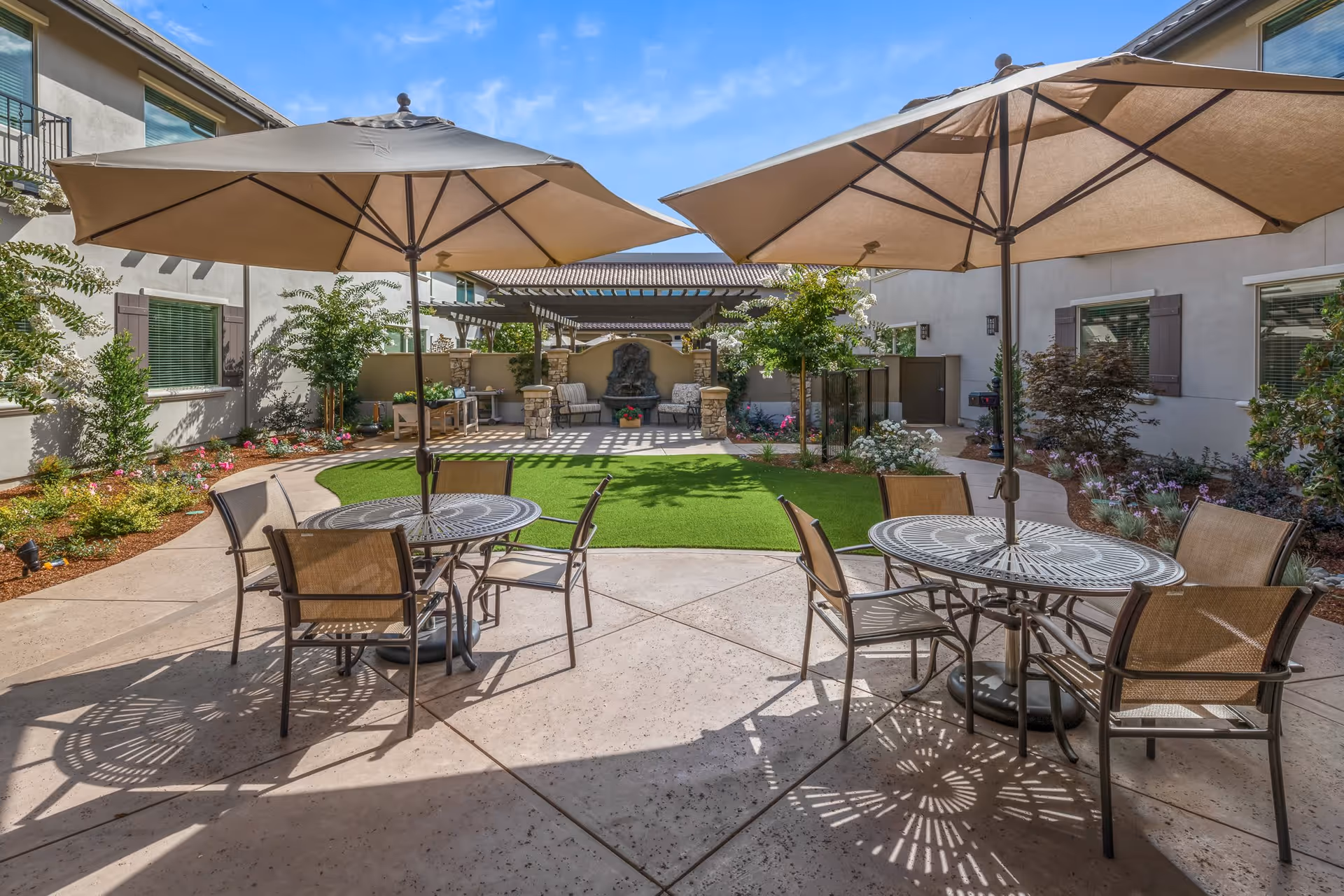Outdoor courtyard area with two round metal tables, each with four chairs and large beige umbrellas providing shade. The courtyard features a green artificial turf lawn, landscaped flower beds, small trees, and a shaded seating area with a water fountain on the far side. The surrounding buildings have windows with shutters and light-colored walls under a clear blue sky.