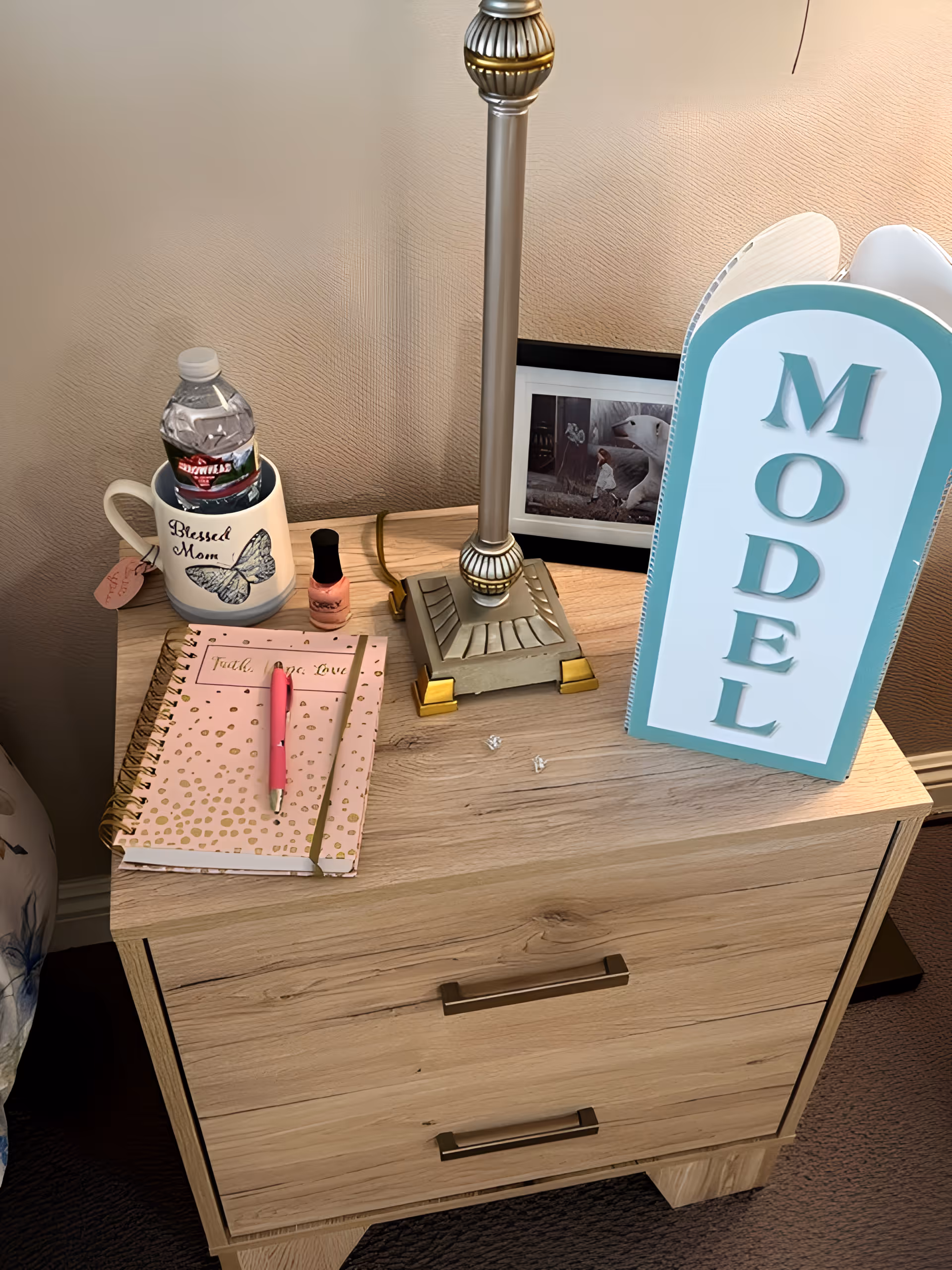 Wooden bedside table topped with a lamp, a water bottle in a 'Blessed Mom' mug, a pink notebook and pen, nail polish, a framed photo, and a decorative sign reading 'MODEL'.