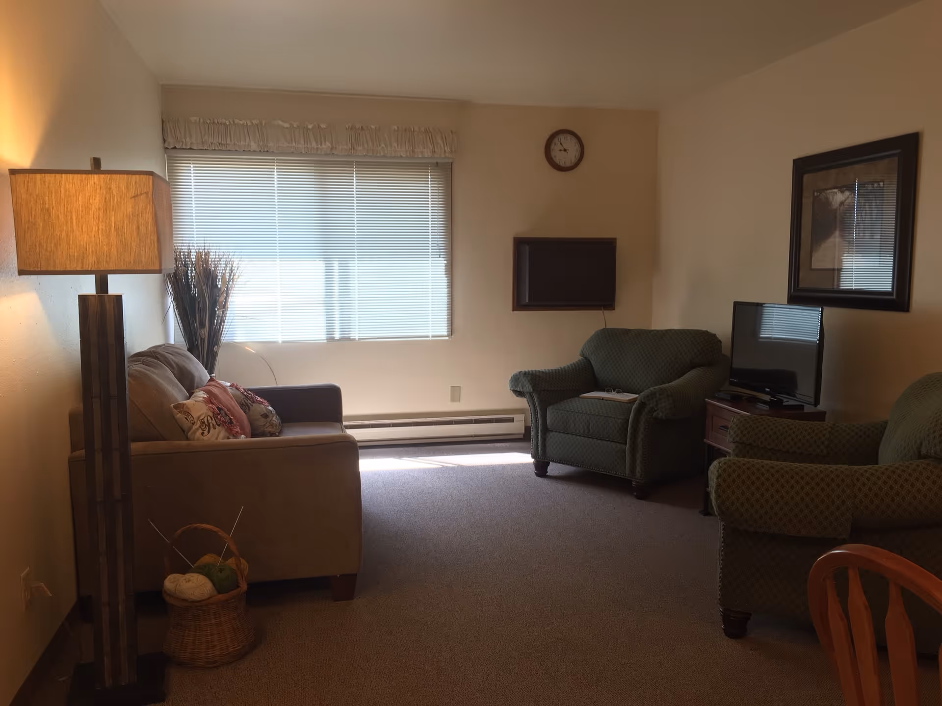 Cozy senior-living living room with beige sofa, green chairs, lamp, and TV.