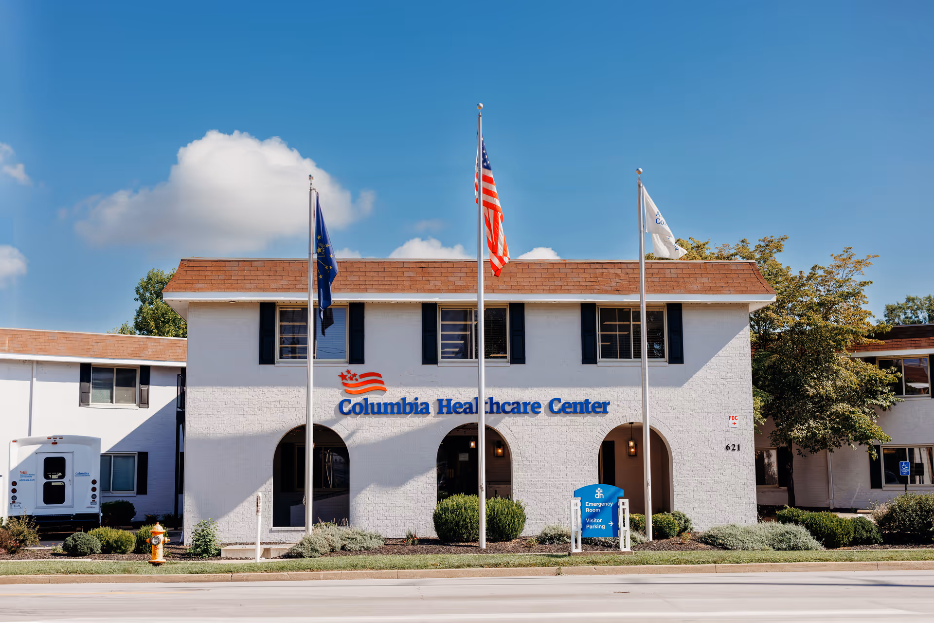 Exterior front view of Columbia Healthcare Center building with three flagpoles displaying flags, a blue sky with some clouds, and landscaping including bushes and a fire hydrant.
