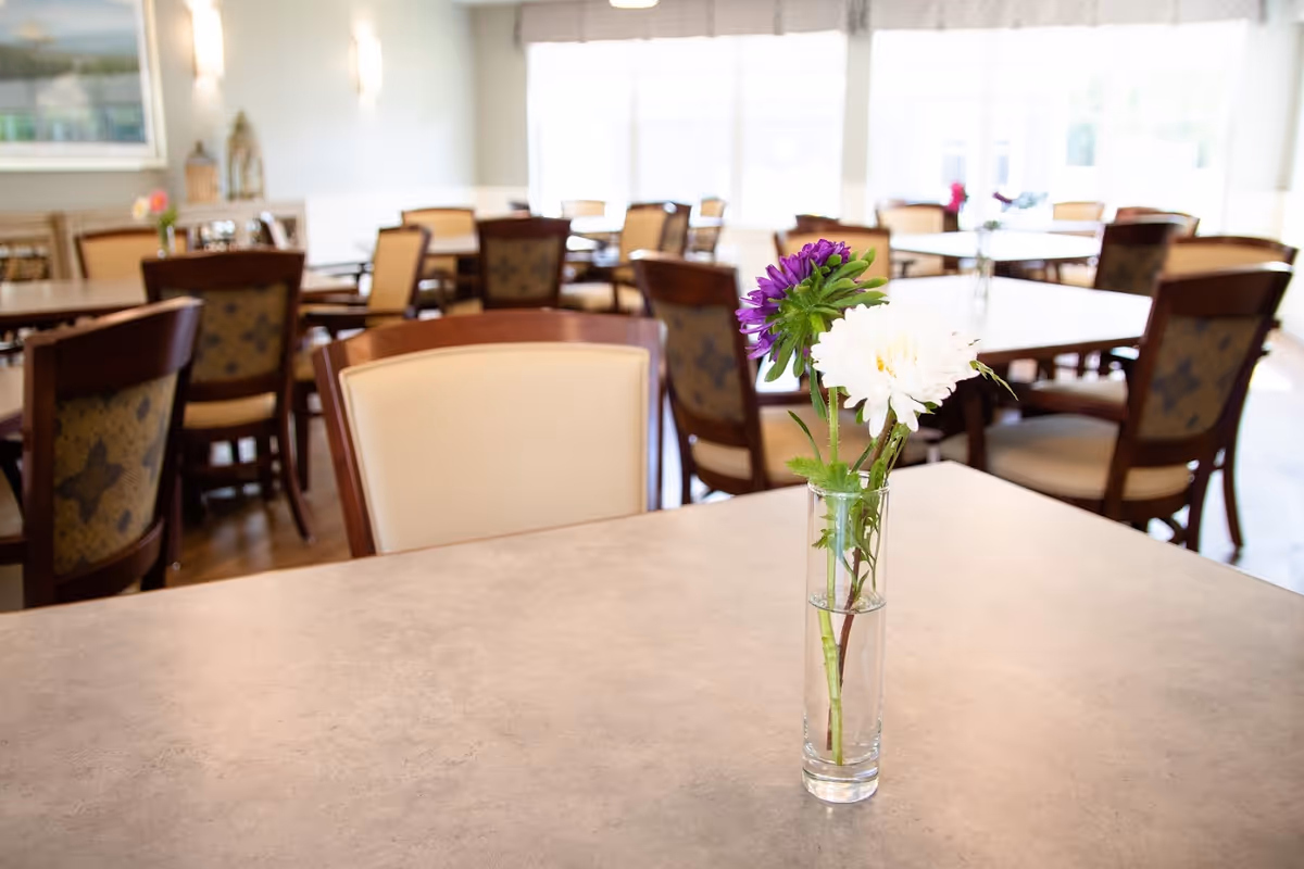 A bright dining room with multiple wooden tables and chairs. A clear glass vase with purple and white flowers is placed on a table in the foreground. Large windows in the background allow natural light to fill the room.