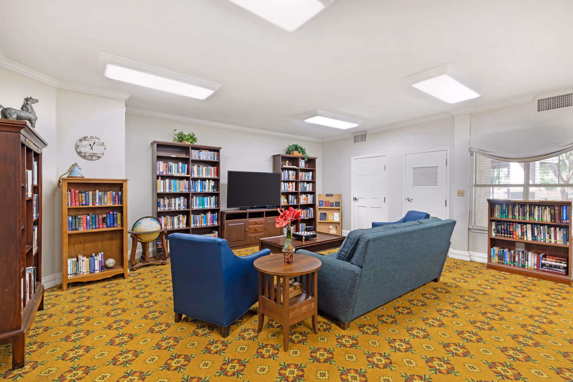 A cozy living room area with a patterned yellow carpet, a blue armchair, a blue sofa, and a wooden side table with a red flower vase. The room features multiple wooden bookshelves filled with books, a globe on a stand, a flat-screen TV on a wooden TV stand, and a window with a light-colored curtain.