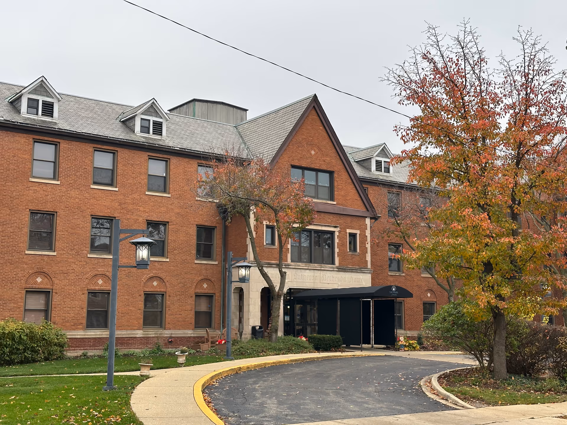 Exterior view of a large brick building with multiple windows and a peaked roof entrance. There is a curved driveway leading to the entrance, which has a black awning with the text 'The Pearl'. Trees with autumn-colored leaves and green shrubs surround the building.