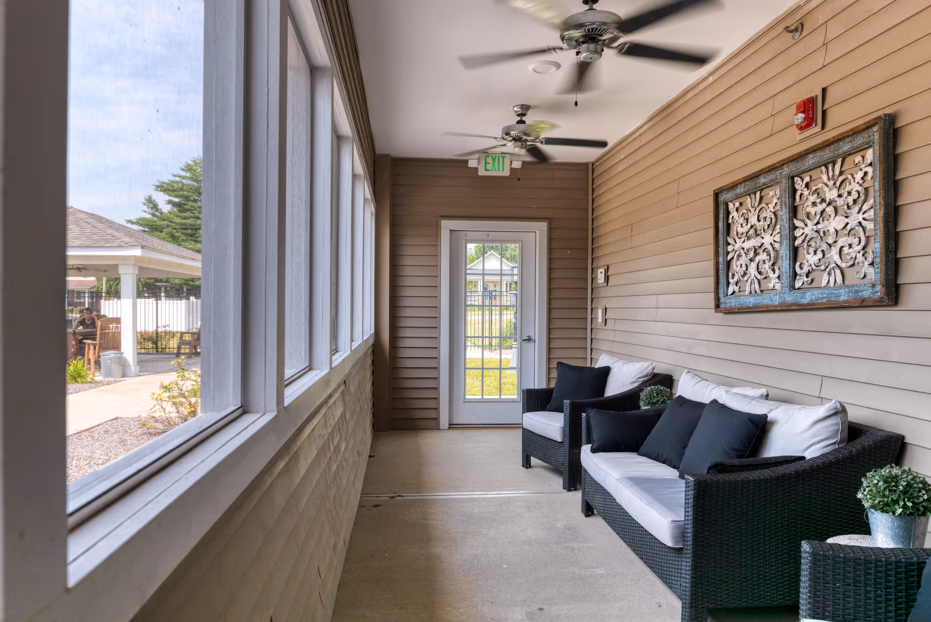 A bright enclosed porch area with large windows on the left side, featuring black wicker furniture with white cushions and black pillows on the right side. The wall behind the furniture is beige with decorative wall art. Ceiling fans are mounted on the white ceiling, and a glass door with an exit sign above it is at the end of the porch. Outside greenery and a gazebo are visible through the windows.