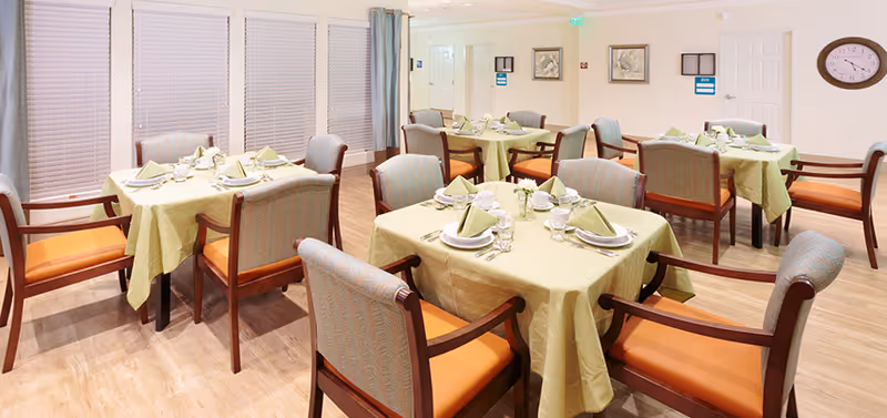 A dining room with several square tables covered with light green tablecloths, each set with plates, napkins, cups, and silverware. The chairs have wooden frames with orange cushions. The room has light-colored walls, large windows with blinds, and a clock on the wall.