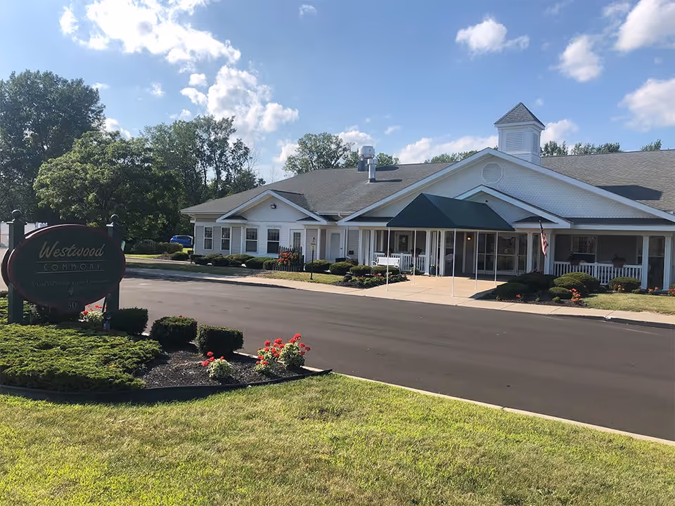 Exterior view of Westwood Commons, a single-story senior living community building with a white facade, green awning over the entrance, and well-maintained landscaping including bushes and flowers. A sign with the facility name is visible on the left side near the driveway.