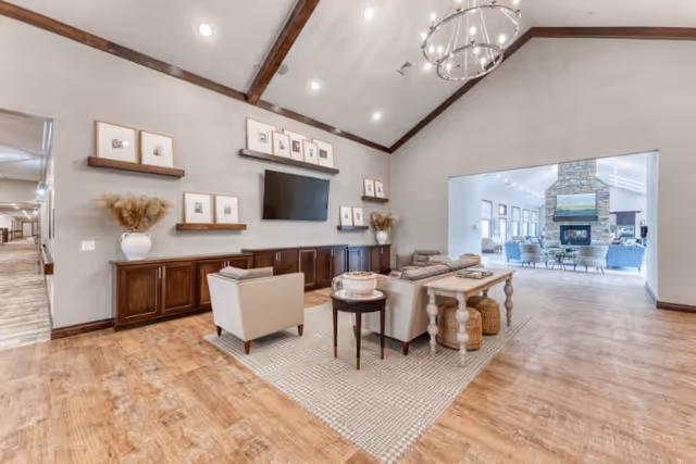Spacious senior living common area with high vaulted ceiling and wooden beams. The room features a seating arrangement with armchairs and a sofa on a large area rug, a wooden console table behind the sofa, and a wall-mounted flat-screen TV above dark wood cabinetry. Decorative framed pictures and vases with dried plants adorn the walls and shelves. Through a large open doorway, another seating area with a stone fireplace and multiple windows is visible.