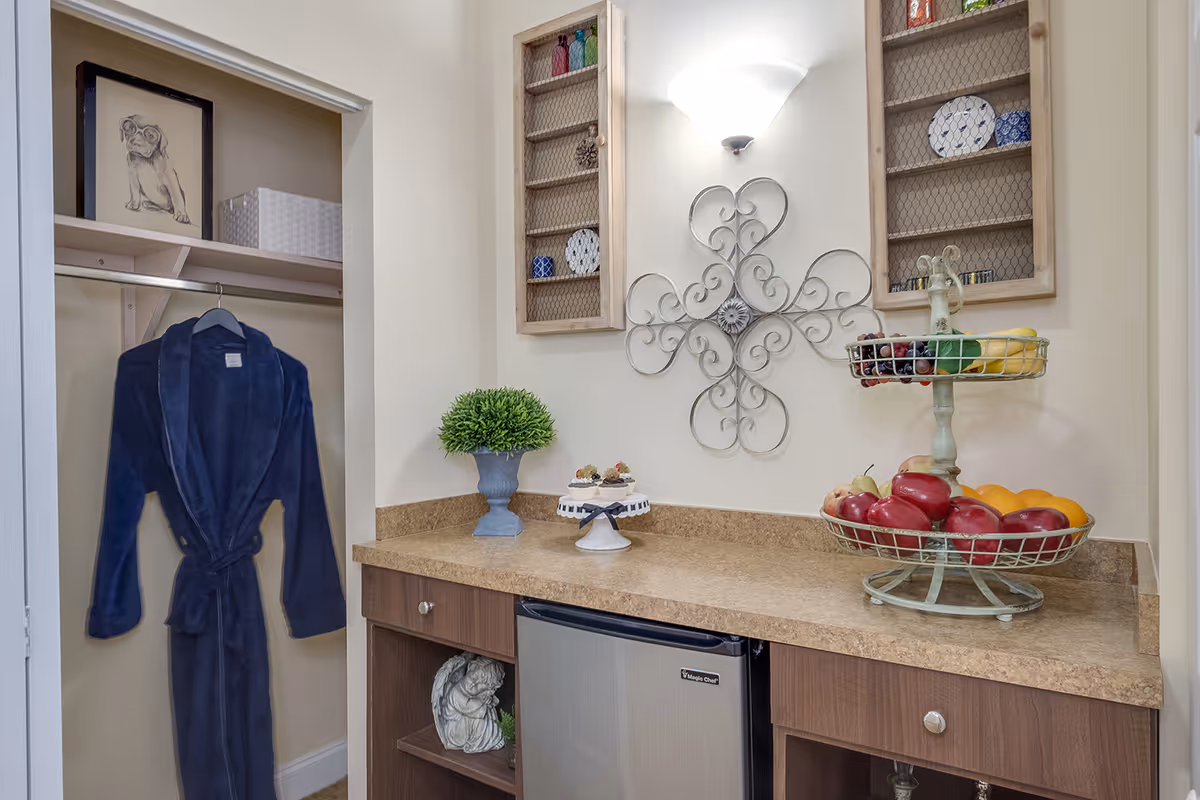 Interior view of a small kitchenette area with a countertop holding a two-tiered fruit basket filled with various fruits, a small potted plant, and a decorative cake stand. Above the countertop is a wall-mounted light fixture and decorative metal wall art. To the left, there is an open closet with a dark blue robe hanging inside and a framed drawing of a dog on the top shelf.