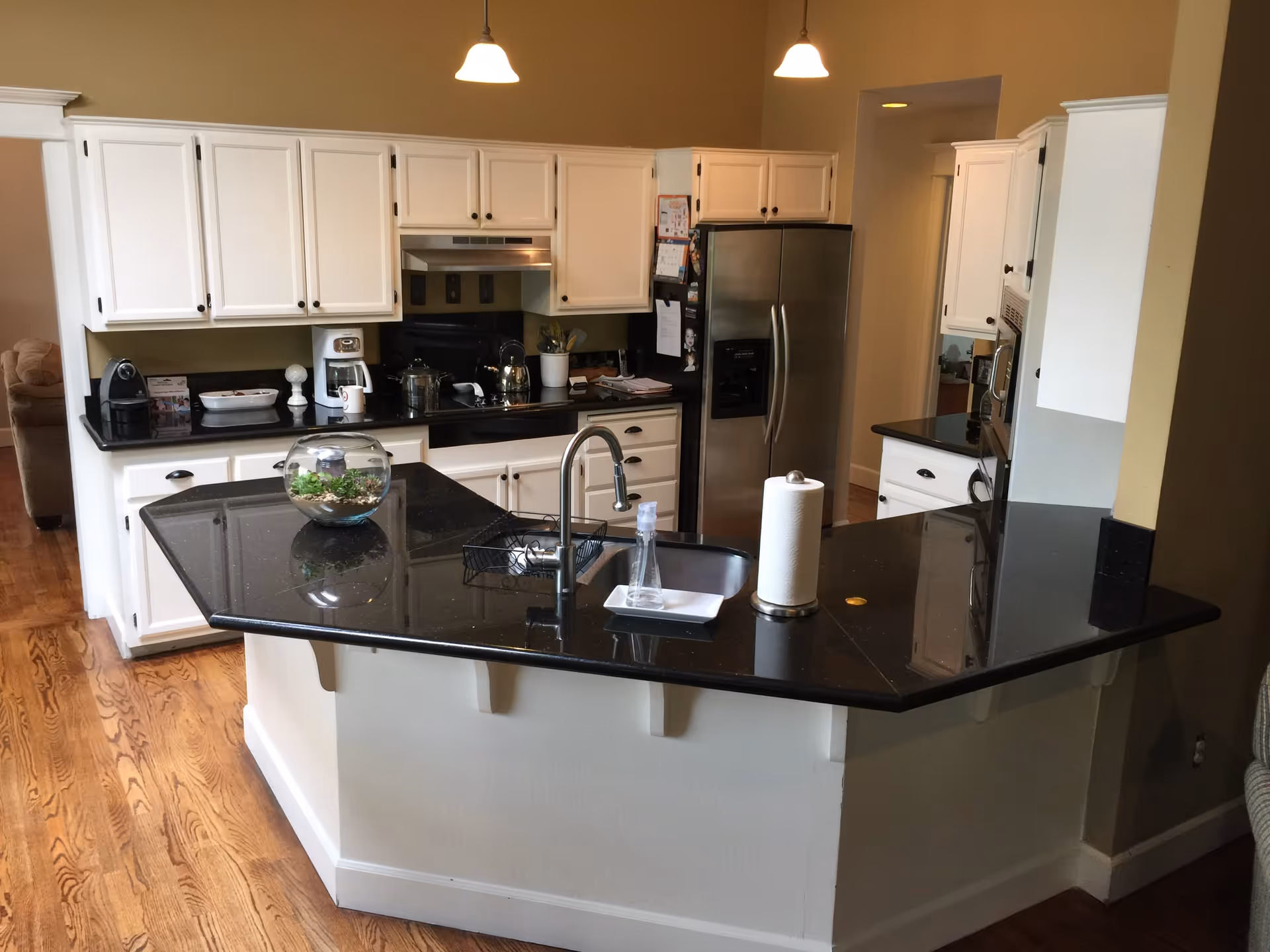 Modern kitchen with white cabinets and black countertops, featuring a central island with a sink, paper towel holder, and a small glass bowl with plants. Stainless steel refrigerator and built-in microwave are visible, along with a coffee maker and other kitchen appliances on the counters. Hardwood floors and pendant lights above the island complete the scene.