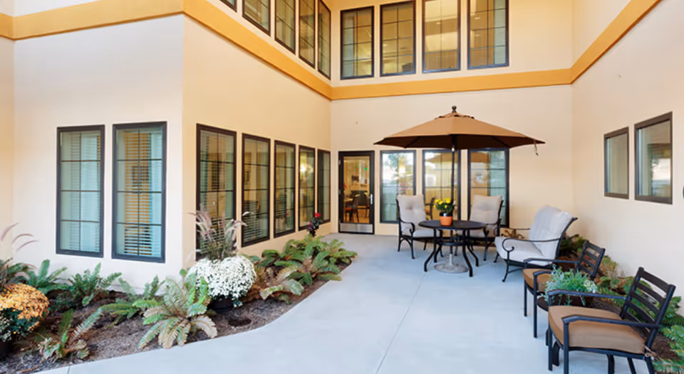 Outdoor courtyard area with a round table and four cushioned chairs under a large brown umbrella. There are additional chairs with cushions along the right side and various plants and flowers along the left side near the building's windows.