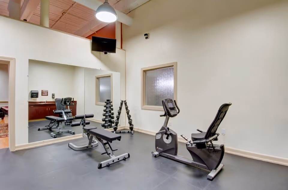 Small fitness room with a recumbent exercise bike, weight bench, dumbbell rack and a large wall mirror.