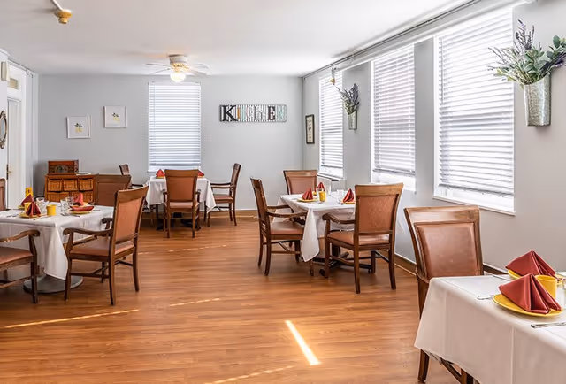 A bright dining room with wooden floors and several tables covered with white tablecloths. Each table is set with red folded napkins, yellow plates, and utensils. The room has multiple windows with white blinds, and decorative wall hangings including a sign that says 'KITCHEN' and flower arrangements in metal wall vases.