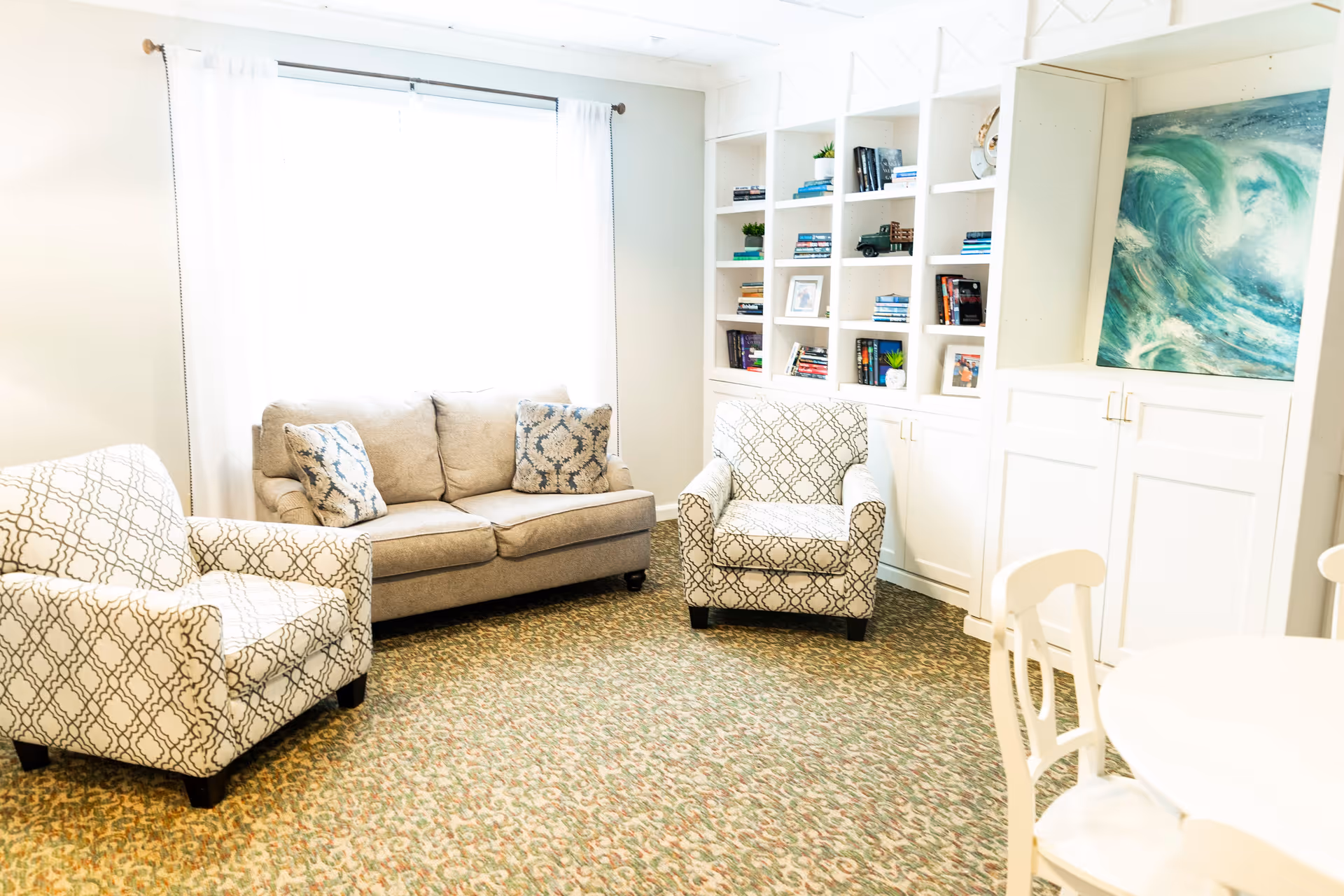 A bright living room with a beige sofa and two patterned armchairs arranged around a carpeted floor. There is a large window with white curtains letting in natural light. Built-in white shelves filled with books and decorative items are on one wall, and a painting of ocean waves is displayed above white cabinets. A round white table with chairs is partially visible in the foreground.