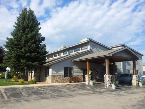 Single-story senior living building with a covered porte-cochère, stone accents, and a large pine tree under a partly cloudy sky.