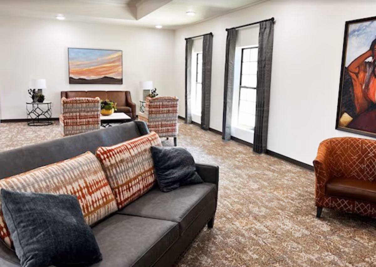 A spacious living room with a gray sofa adorned with patterned cushions and dark pillows in the foreground. Behind the sofa, there are two patterned armchairs facing a brown leather couch with a coffee table in between. The room has large windows with dark curtains, a patterned carpet, and two side tables with lamps. A colorful painting hangs on the wall to the right.