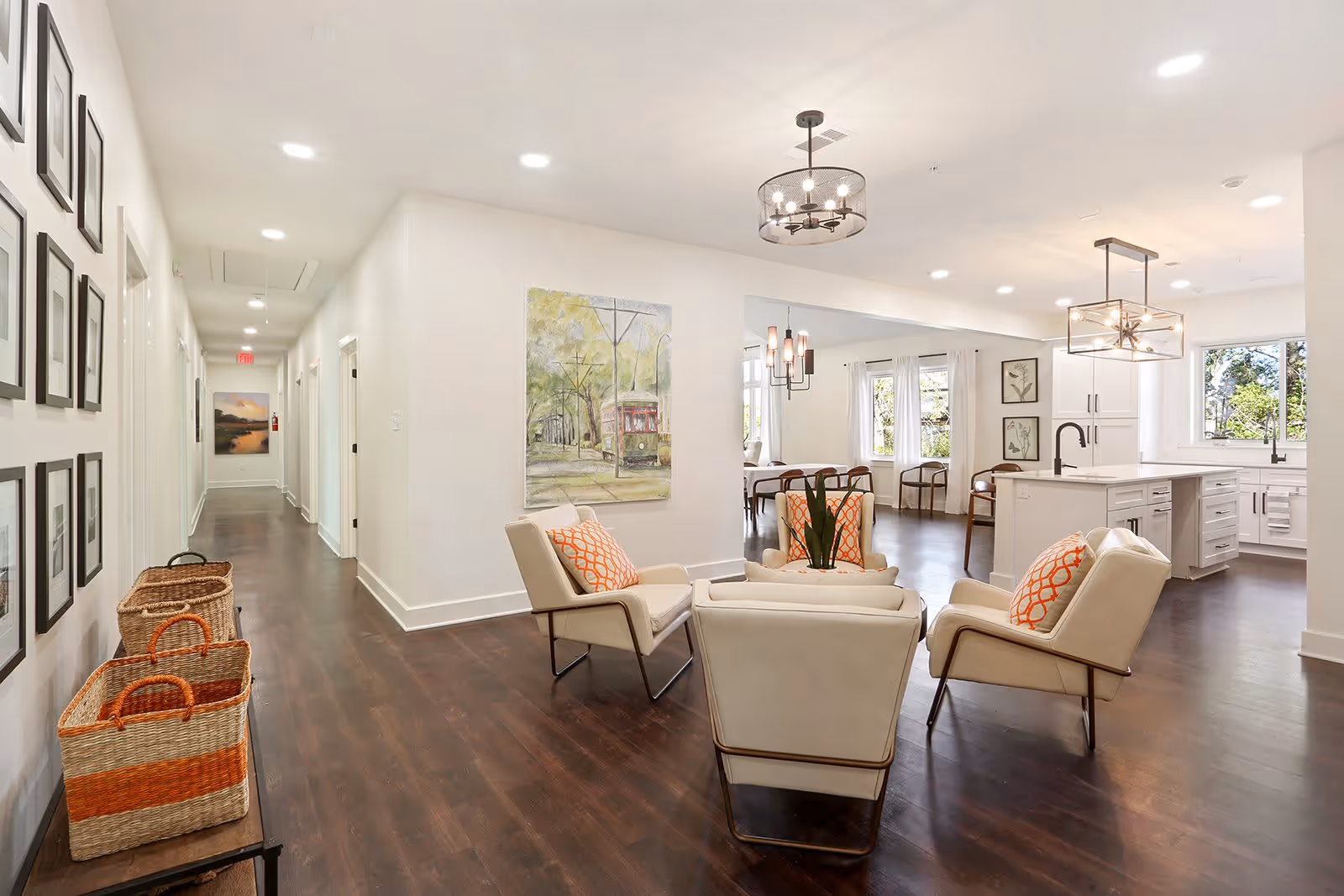 A bright and modern interior space featuring a seating area with four cream-colored armchairs with orange patterned pillows arranged around a small table. The room has dark wooden floors and white walls adorned with framed artwork. In the background, there is a kitchen area with white cabinetry, a large island with a sink, and pendant lighting. A hallway with additional framed pictures and baskets on a bench extends to the left.