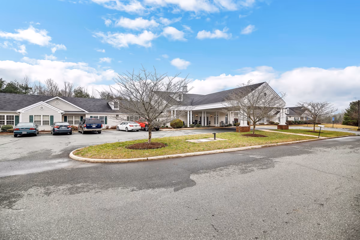 Exterior view of Spring Oak at Louisa facility showing a single-story building with a covered entrance, several parked cars, leafless trees, and a partly cloudy sky.