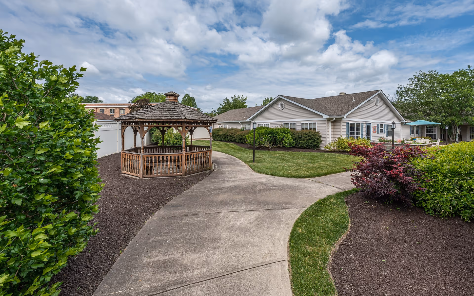 Outdoor garden area at Arden Courts - ProMedica Memory Care Community in Wilmington featuring a wooden gazebo, paved walking paths, well-maintained green lawns, bushes, and trees under a partly cloudy sky.