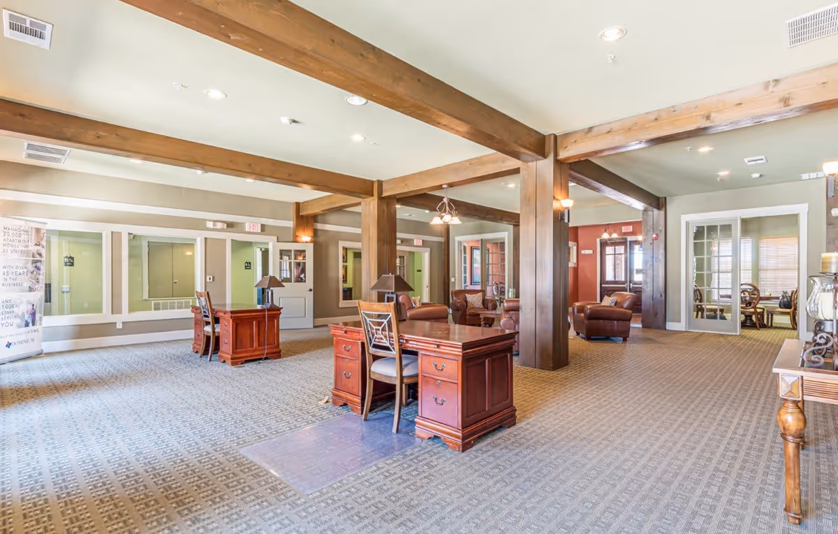 A spacious interior common area with wooden beams on the ceiling, carpeted floor, and several wooden desks with chairs and lamps. There are leather armchairs and sofas in the background near glass doors and windows, creating a welcoming lounge space.