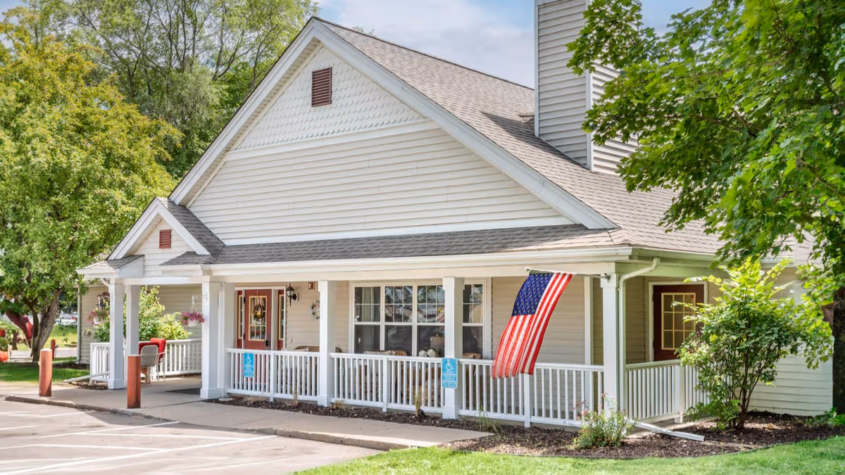 Exterior view of a single-story building with beige siding and a gabled roof, featuring a covered porch with white railings and an American flag hanging near the entrance. The building is surrounded by green trees and landscaping, with a parking area in front.