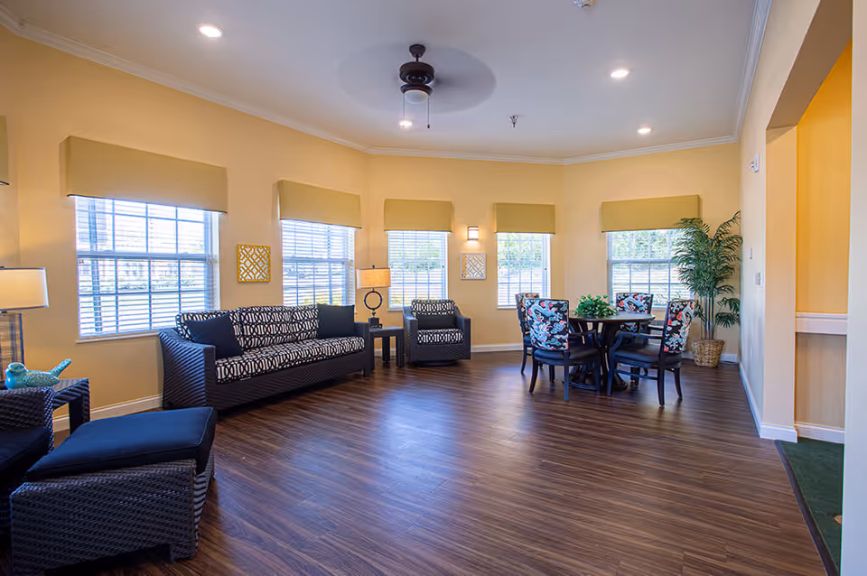 Bright communal living room with wicker sofas, a round dining table with floral chairs, multiple windows, and wood floors.